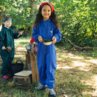 A girl in a blue Originals Waterproof Puddle Suit smiles, holding a dish with greenery, whilst outdoors. Another child stands behind her.